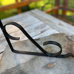 May include: A black, wrought iron shelf bracket with a decorative scroll design. The bracket is angled and has a flat base for mounting. The scroll adds a touch of elegance to the functional piece.
