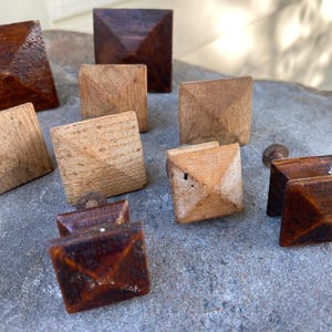 May include: A collection of square wooden knobs in various shades of brown and tan. The knobs have a pyramid-like design on the front and are likely for furniture or decorative purposes. Some knobs show signs of wear, adding to their rustic charm.