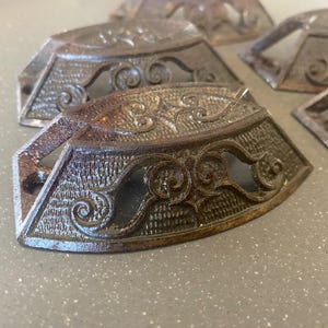 May include: A collection of antique, ornate metal drawer pulls. The pulls have a dark, aged finish with intricate scrollwork and a textured surface. The design includes a central, decorative element and mounting holes for installation. The hardware is likely for furniture restoration.