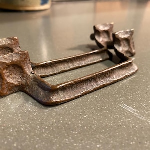 May include: A handcrafted, bronze-colored metal object with a textured surface. The piece has a rectangular base with two parallel bars and two decorative, raised elements. The metal has a rustic, aged appearance.