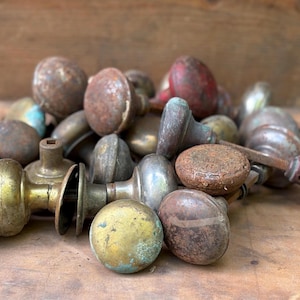 May include: A collection of vintage door knobs in various finishes including tarnished brass, rusted iron, and weathered metal. The knobs feature round, spherical shapes with some showing signs of oxidation, patina, and wear, scattered in a pile.