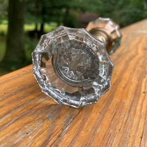 May include: A clear, faceted glass door knob with a multi-sided, geometric shape and a starburst pattern at the center. The knob features a metallic base that connects to a cylindrical shaft, showcasing a reflective, transparent design.