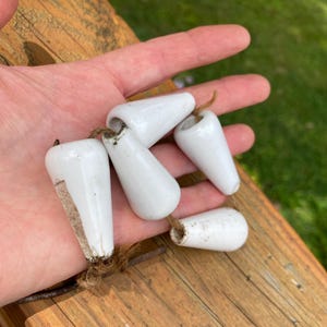 May include: A collection of six white ceramic drawer pulls with a teardrop shape. Each pull is attached to a length of natural twine. The pulls are held in a hand, with a wooden deck and green grass in the background.