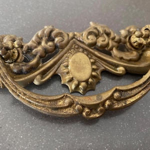 May include: Ornate antique brass drawer pull with a curved handle and decorative floral and scrollwork details. The center features an oval medallion with a sunburst design. A vintage-style hardware piece.