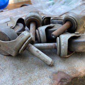 May include: A collection of vintage metal casters, featuring a wheel, bracket, and stem. The casters are primarily silver with signs of rust, indicating age and use. The wheels are round and appear to be made of a hard material.