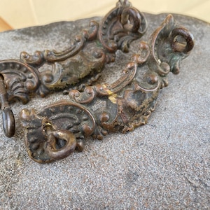 May include: Antique bronze-colored drawer pulls with ornate, scrollwork designs. The hardware includes a screw and a loop for attaching to a drawer. The pulls have a weathered appearance, suggesting age and use. Ideal for vintage furniture restoration.