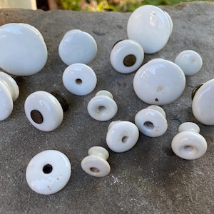 May include: A collection of various white ceramic knobs of different sizes and shapes. Some knobs feature a smooth, rounded top, while others have a central hole or a metallic base attachment. The knobs are arranged in a group, displaying a glossy finish.