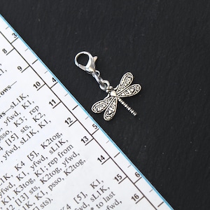 Könnte beinhalten: Silberner Libellen-Anhänger mit detaillierten Flügeln, befestigt an einem Verschluss. Der Anhänger befindet sich vor einem schwarzen Hintergrund mit einem Teil eines Lineals und Strickmusters.