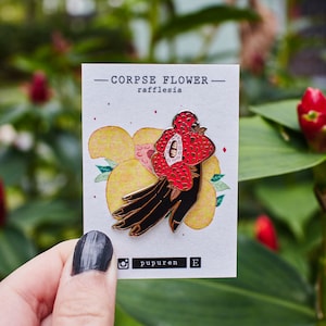 Op de afbeelding: Een zwarte en rode emaille pin met een hand die een rode bloem vasthoudt met de tekst "CORPSE FLOWER - rafflesia -" en de Instagram-handle "pupuren" op een witte kaart.