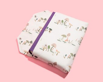 May include: A white book cover with a purple book inside. The cover features a repeating pattern of mushrooms and trees in shades of brown, green, and orange.