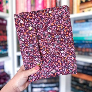 May include: A rectangular fabric book sleeve featuring a dark burgundy background with a dense pattern of small pink, orange, and white flowers interspersed with white human skulls and bones. The sleeve has a fold-over flap secured by two gold snaps.