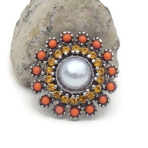May include: A silver brooch with a large pearl in the center surrounded by orange and yellow rhinestones.
