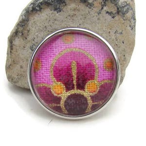 May include: A round, silver-toned button with a pink and gold fabric design. The fabric has a geometric pattern with a central flower shape.
