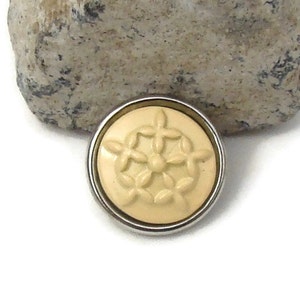 May include: A round, cream-colored button with a silver-colored metal rim. The button features a raised floral design in the center. The button is set against a neutral background.