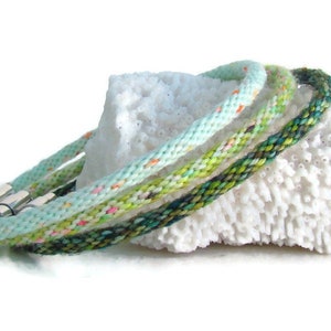May include: Three braided bracelets in shades of green, blue, and white with silver clasps. The bracelets are displayed on a white coral-like surface.