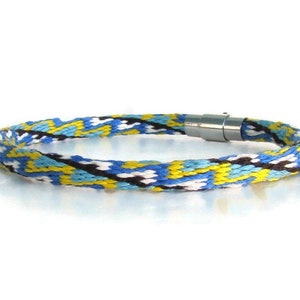May include: Braided bracelet with a geometric pattern in blue, yellow, black, and white. The bracelet has a silver-colored magnetic clasp. The woven design creates a textured appearance, suitable for everyday wear.