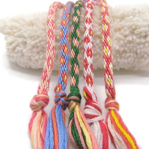 May include: Close-up of six colorful friendship bracelets with woven patterns. The bracelets feature pink, blue, green, and red threads, with knotted ends and tassels. The background is a white, textured surface.