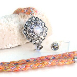 May include: A colorful braided lanyard with a silver retractable badge holder and a silver and white decorative charm. The charm features a pearl-like center and a floral design.