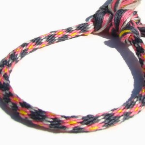 May include: A woven bracelet featuring a repeating pattern of pink, yellow, white, and dark gray threads. The bracelet is knotted at the end with matching threads, creating a loop. The bracelet is approximately 7 inches in circumference.
