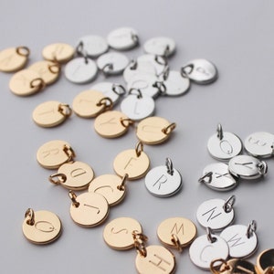 Könnte beinhalten: Eine Sammlung von goldenen und silbernen Metallanhängern mit eingravierten Buchstaben des Alphabets. Die Anhänger sind auf einer weißen Oberfläche angeordnet.