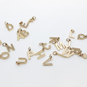 Puede incluir: Una colección de abalorios de letras de metal dorado, esparcidos sobre una superficie blanca. Las letras incluyen A, B, C, D, E, F, H, J, L, M, N, O, P, Q, U, V, W y Z.