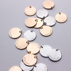 Może przedstawiać: Zbiór okrągłych metalowych zawieszek z wygrawerowanymi na nich inspirującymi słowami. Zawieszki są w kolorze srebrnym i złotym i mają otwór do przymocowania do naszyjnika lub bransoletki. Słowa obejmują "powerful", "loved", "resilient", "worthy", "fearless", "present", "smart", "creative", "strong", "grateful".