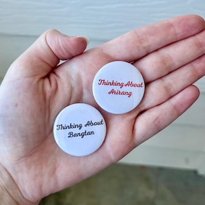 May include: Two white round buttons with black text that reads "Thinking About Bangtan" and "Thinking About Arirang" in red. The buttons are held in an open palm.