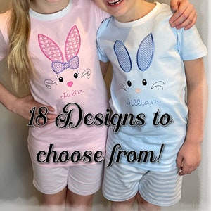 May include: Two children wearing matching pajamas with bunny embroidery. The girl's pajamas are pink with a pink bunny and the boy's pajamas are blue with a blue bunny. The pajamas are personalized with the names "Julia" and "William". 18 Designs to choose from!