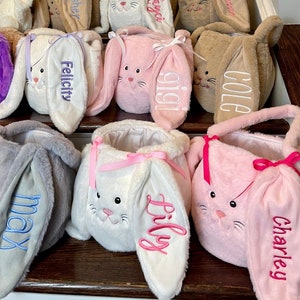 May include: Ten personalized Easter baskets with bunny ears. The baskets are made of soft, plush fabric and come in a variety of colors, including pink, white, gray, and brown. Each basket has a different name embroidered on it, including "Max", "Lily", "Charlie", "Cole", "Gigi", "Felicity", "Asher", "Keira", "Gwen", and "Vera".