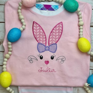 K&ouml;nnte beinhalten: Rosa Langarmshirt und gestreifte Hose mit einem gestickten Hasengesicht-Design. Der Hase hat rosa Ohren mit Muster, eine lila Schleife und den Namen "Julia" darunter gestickt. Eine Holzperlen-Girlande mit bunten Ostereiern umgibt das Set.