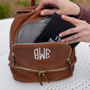 May include: A brown faux leather backpack with gold hardware and a white monogram "BWE" on the front. The backpack is open and a black book with the text "The Secret" is partially visible inside.