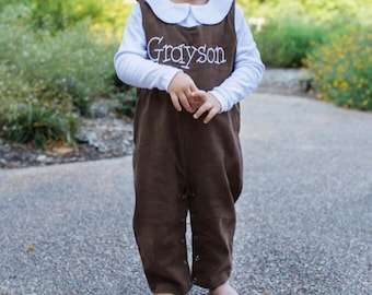 May include: A young child wearing a brown overall with a white collared shirt and the name "Grayson" embroidered on the front. The child is standing on a paved path.