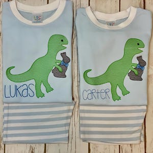 K&ouml;nnte beinhalten: Zwei hellblaue Schlafanz&uuml;ge mit wei&szlig;em Besatz, mit einem gr&uuml;nen Dinosaurier, der einen braunen Hasen mit einer blauen Schleife h&auml;lt. Ein Set hat den Namen "Lukas" in Blau gestickt, das andere "Carter". Die Schlafanzughosen haben blau-wei&szlig;e Streifen.