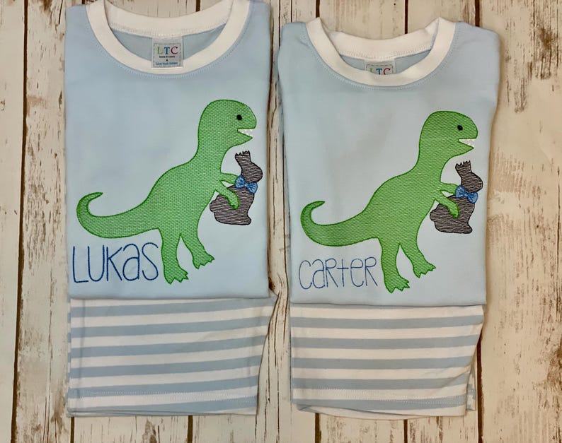 K&ouml;nnte beinhalten: Zwei hellblaue Schlafanz&uuml;ge mit wei&szlig;em Besatz, mit einem gr&uuml;nen Dinosaurier, der einen braunen Hasen mit einer blauen Schleife h&auml;lt. Ein Set hat den Namen "Lukas" in Blau gestickt, das andere "Carter". Die Schlafanzughosen haben blau-wei&szlig;e Streifen.