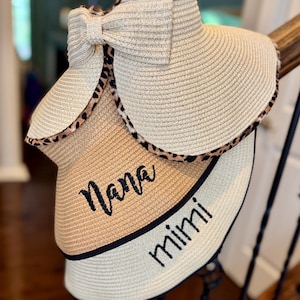 May include: Three straw visors with a bow and leopard print trim. One visor has "Nana" embroidered in black, and another has "Mimi". The visors are designed to shield the face from the sun.