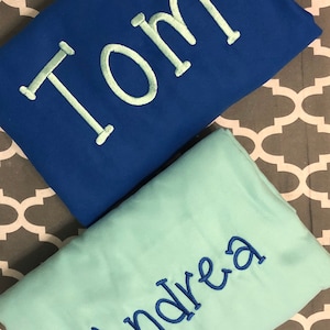 May include: Two personalized blankets, one blue with the name "Tom" embroidered in white, and one light blue with the name "Andrea" embroidered in blue.