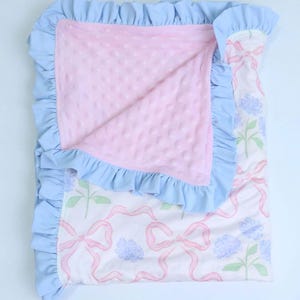 Puede incluir: Una manta para beb&eacute; con un borde con volantes azul claro. La manta tiene un centro rosa texturizado y un exterior estampado con lazos rosas, flores azules y tallos verdes. La manta est&aacute; doblada.