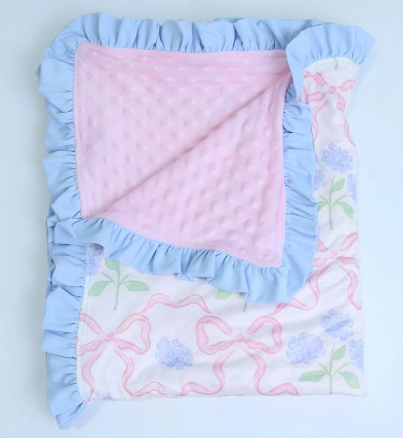 Puede incluir: Una manta para beb&eacute; con un borde con volantes azul claro. La manta tiene un centro rosa texturizado y un exterior estampado con lazos rosas, flores azules y tallos verdes. La manta est&aacute; doblada.