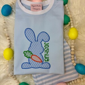 May include: A light blue t-shirt with a white trim around the neck. The shirt features an embroidered bunny holding a carrot with the name "Joshua" embroidered below. The shirt is surrounded by pastel colored Easter eggs.