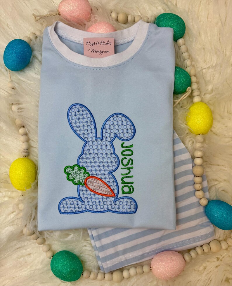 May include: A light blue t-shirt with a white trim around the neck. The shirt features an embroidered bunny holding a carrot with the name "Joshua" embroidered below. The shirt is surrounded by pastel colored Easter eggs.