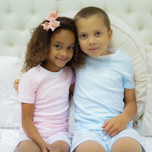 May include: Two children, a girl with curly brown hair wearing a pink shirt with the word "Lola" embroidered on it and a boy wearing a light blue shirt with the word "Kaiden" embroidered on it, are sitting on a white bed with white pillows.