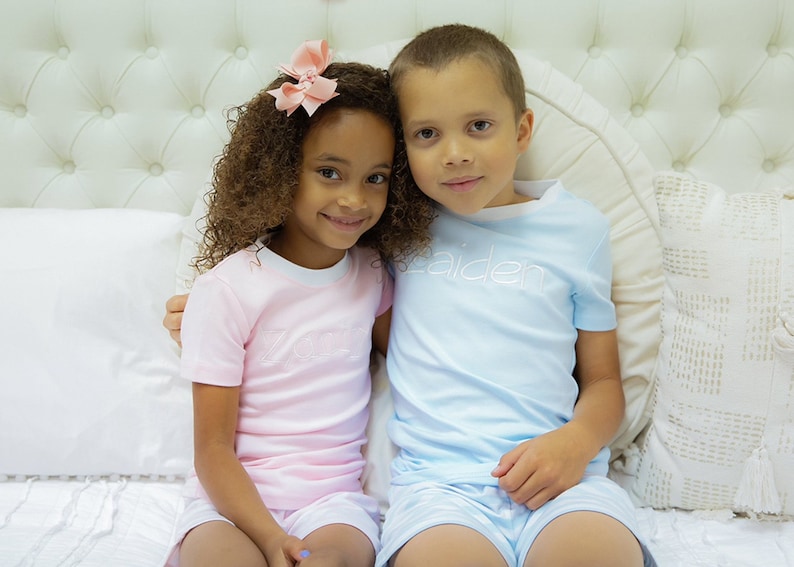 May include: Two children, a girl with curly brown hair wearing a pink shirt with the word "Lola" embroidered on it and a boy wearing a light blue shirt with the word "Kaiden" embroidered on it, are sitting on a white bed with white pillows.
