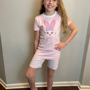 May include: A pink short-sleeved shirt with a white trim and an embroidered bunny with a purple bow. The shirt says "Julia" on the front.