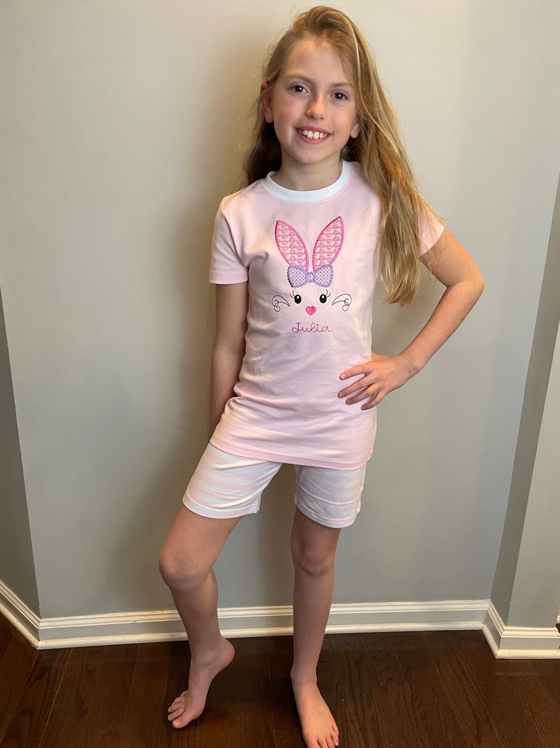 May include: A pink short-sleeved shirt with a white trim and an embroidered bunny with a purple bow. The shirt says "Julia" on the front.