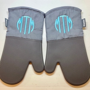 May include: A pair of gray oven mitts with a dark gray silicone grip. Each mitt features a light blue embroidered monogram within a circle. The brand name "Honla" is printed on a small tag on the side.