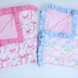 Puede incluir: Dos mantas para beb&eacute;, una rosa y otra azul, con bordes con volantes. La manta rosa presenta un patr&oacute;n de lazo, mientras que la azul tiene un dise&ntilde;o floral. Ambas tienen un centro suave y texturizado.
