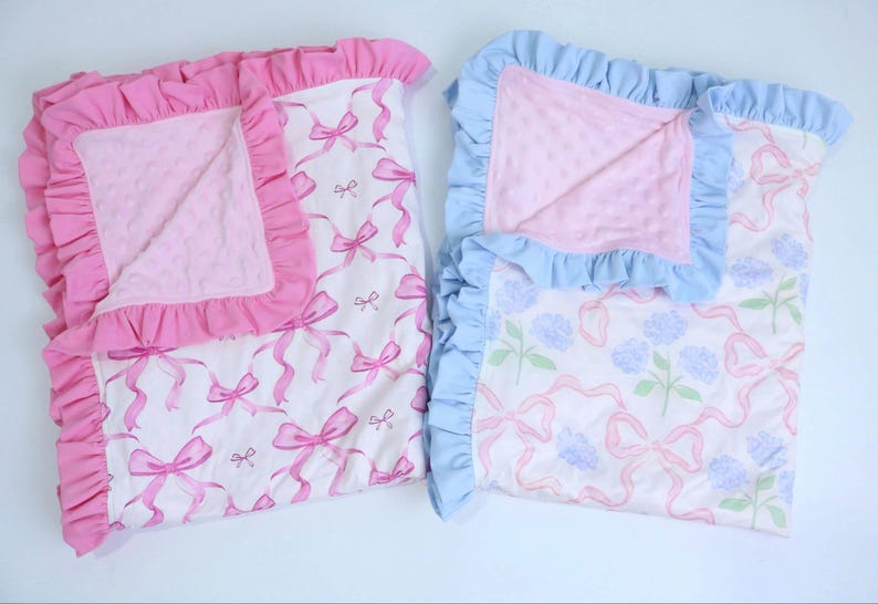 Puede incluir: Dos mantas para beb&eacute;, una rosa y otra azul, con bordes con volantes. La manta rosa presenta un patr&oacute;n de lazo, mientras que la azul tiene un dise&ntilde;o floral. Ambas tienen un centro suave y texturizado.