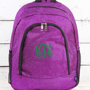 May include: A purple glitter backpack with black trim and zippers. The backpack has a green monogram on the front. The bag has a mesh side pocket and a top handle. The background is a white wooden surface.