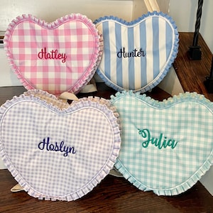 May include: Four heart-shaped pillows with ruffled edges. One is pink gingham with "Hatley" embroidered in pink. Another is blue and white striped with "Hunter" in blue. The third is lavender gingham with "Hoslyn" in blue. The last is aqua gingham with "Julia" in green.