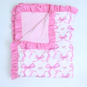 Puede incluir: Una manta rosa para beb&eacute; con fondo blanco y estampado de lazos rosas. La manta tiene un borde rosa con volantes y un interior rosa suave y texturizado. Dise&ntilde;ada para beb&eacute;s y ni&ntilde;os peque&ntilde;os.